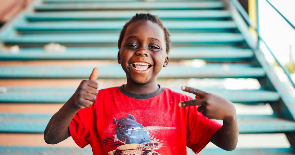 Young boy wearing a red cars tshirt smiling and giving a thumbs up and sitting on stairs outside.