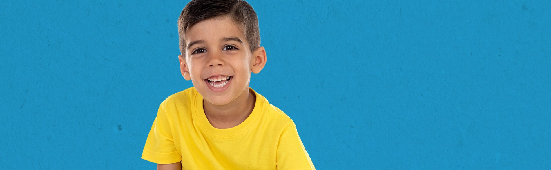 A boy smiling at the camera wearing a yellow tshirt in front of a solid blue background.