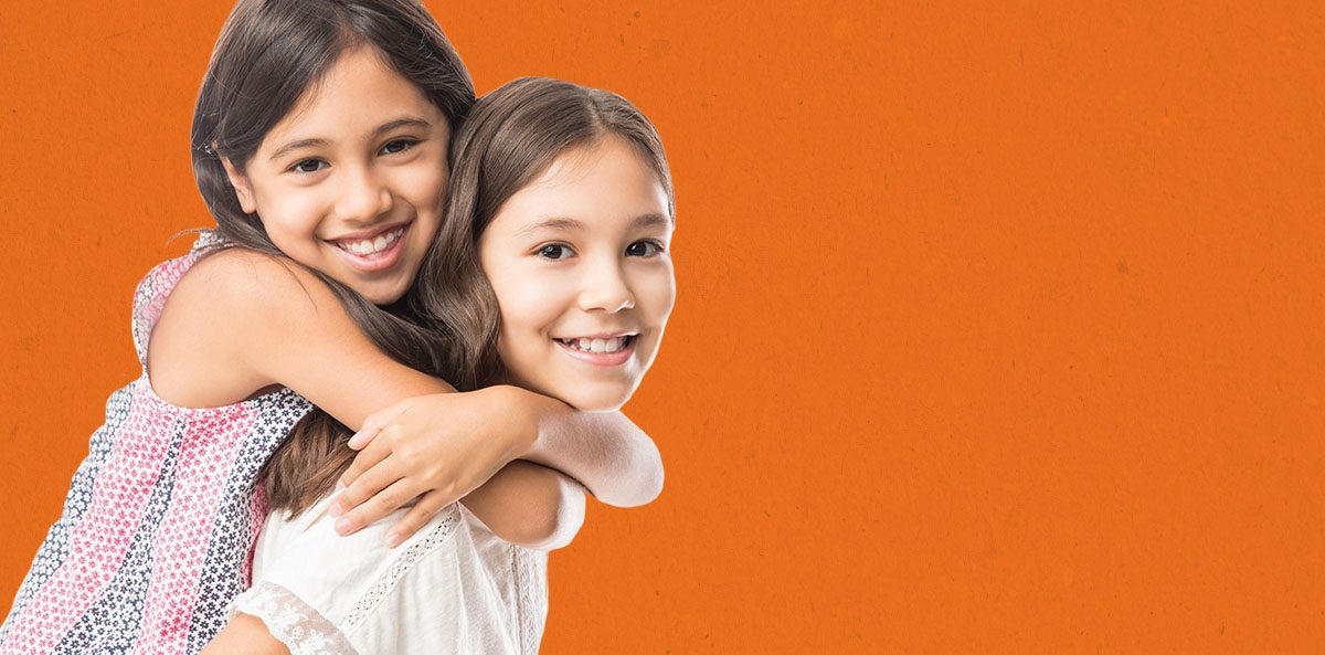 Two girls hugging and smiling at the camera in front of a solid orange background.