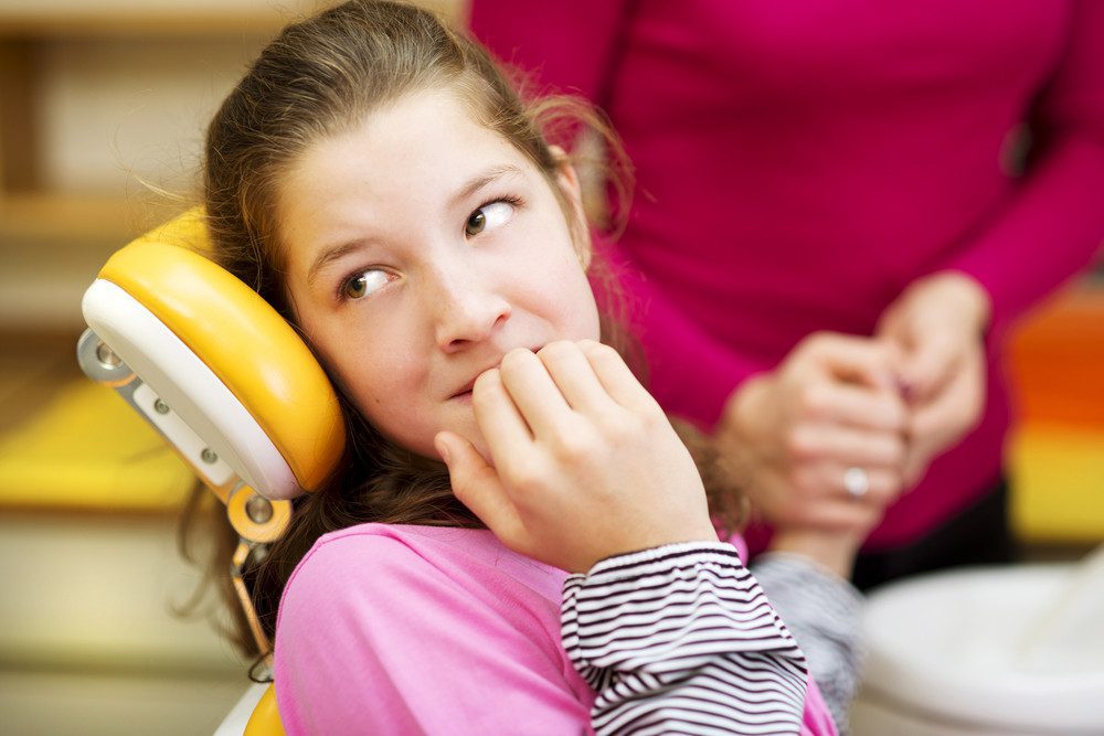 Young girl looking scared sitting in a dentist chair while a woman holds her hand in the background.